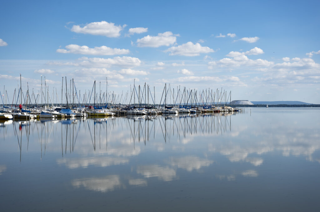In Mardorf liegen zahlreiche Segelboote an einem Steg. Kleine Wolken am strahlend blauen Himmel spiegeln sich im Steinhuder Meer.