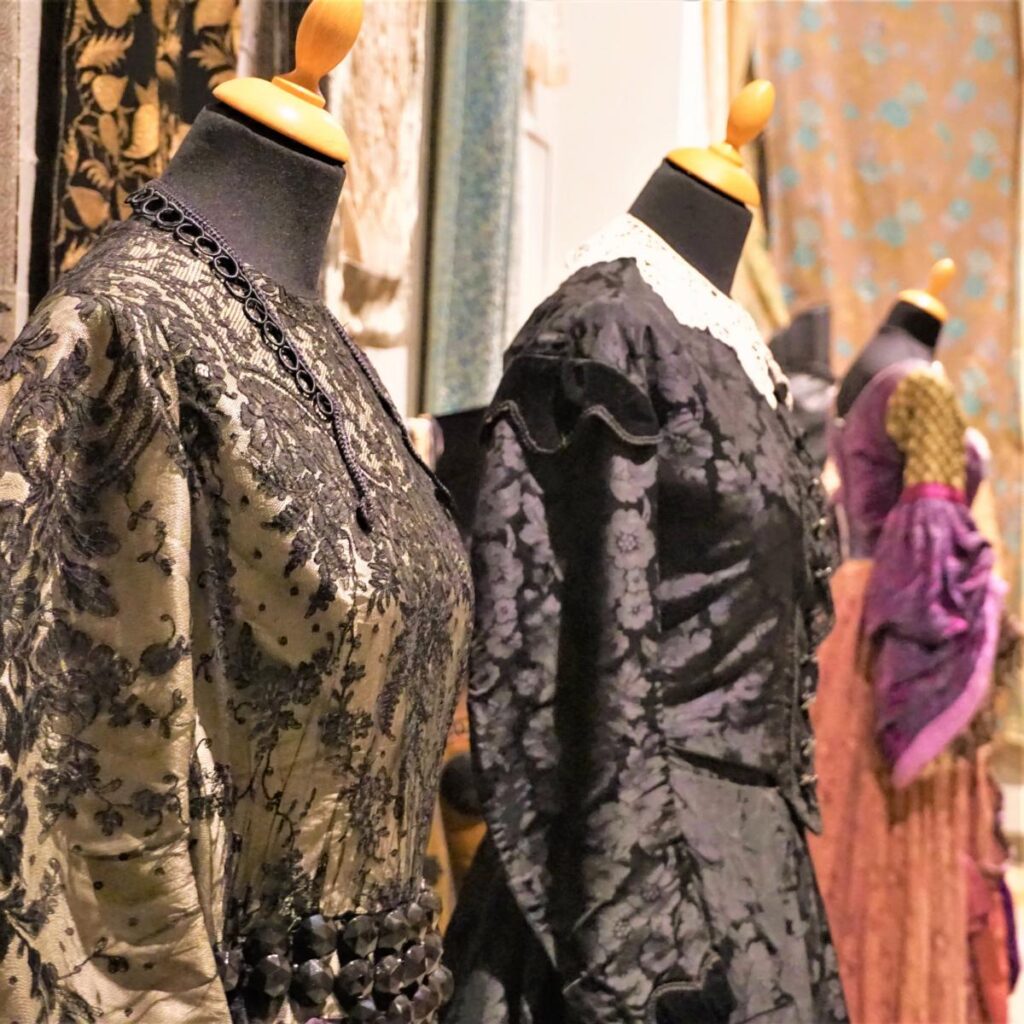 Historische Kleider auf Schneiderpuppen mit Spitze, Stickereien und Mustern in dunklen Farben, ausgestellt in einem Museum.