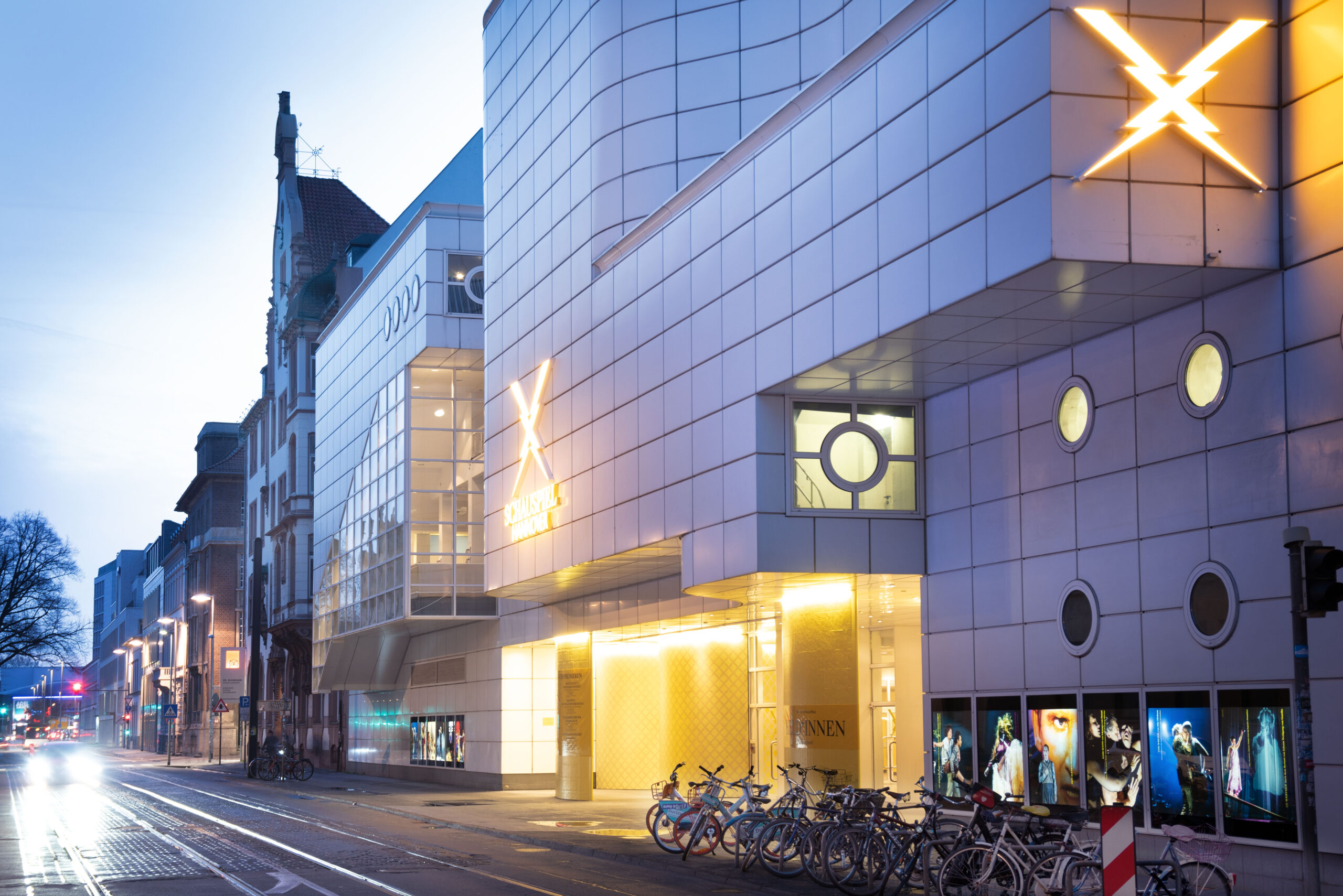 Beleuchteter Eingang des Schauspielhauses Hannover mit moderner Fassade und Fahrrädern am Straßenrand.