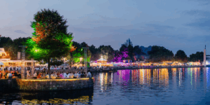 Wieczorny festiwal nad jeziorem z kolorowymi światłami, mnóstwem ludzi i klimatycznymi odbiciami w wodzie.