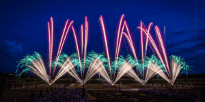 Fogos de artifício coloridos sobre os jardins barrocos à noite, vistos por uma grande multidão.