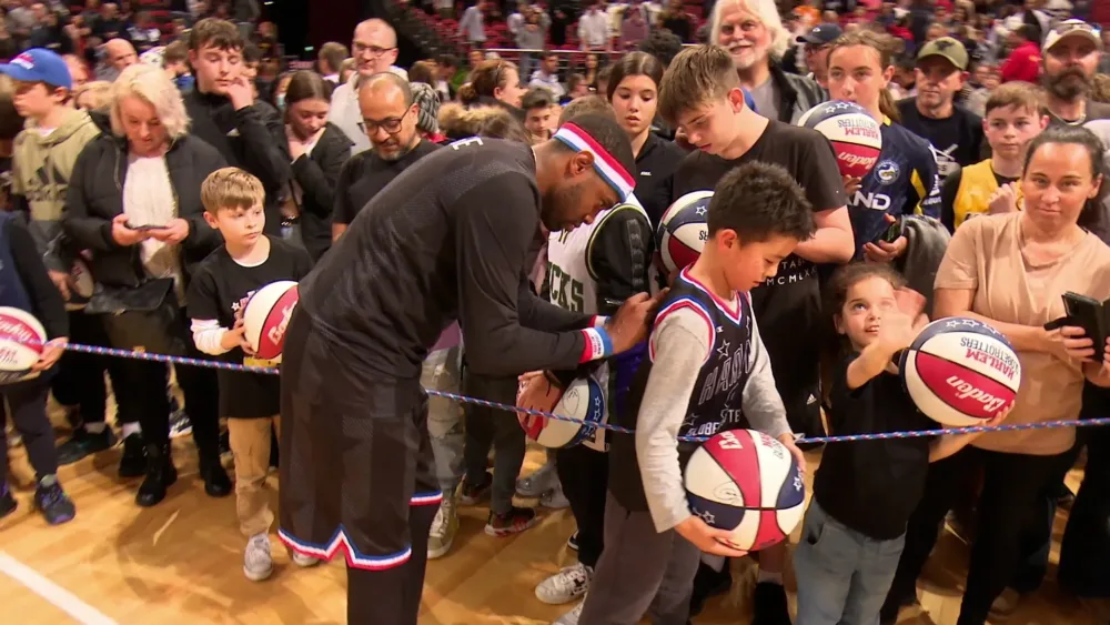 Ein Basketballspieler unterschreibt einem Kind ein Trikot; viele Fans mit Bällen warten dahinter.
