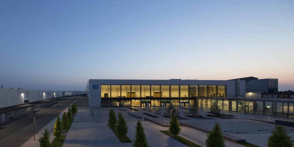 Moderne Messehalle 19 in Hannover bei Abenddämmerung mit beleuchtetem Innenraum und leerem Vorplatz.