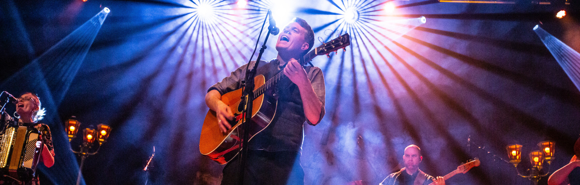 Mehrköpfige Band auf einer bunt beleuchteten Bühne, der Sänger spielt Gitarre und singt leidenschaftlich ins Mikrofon.