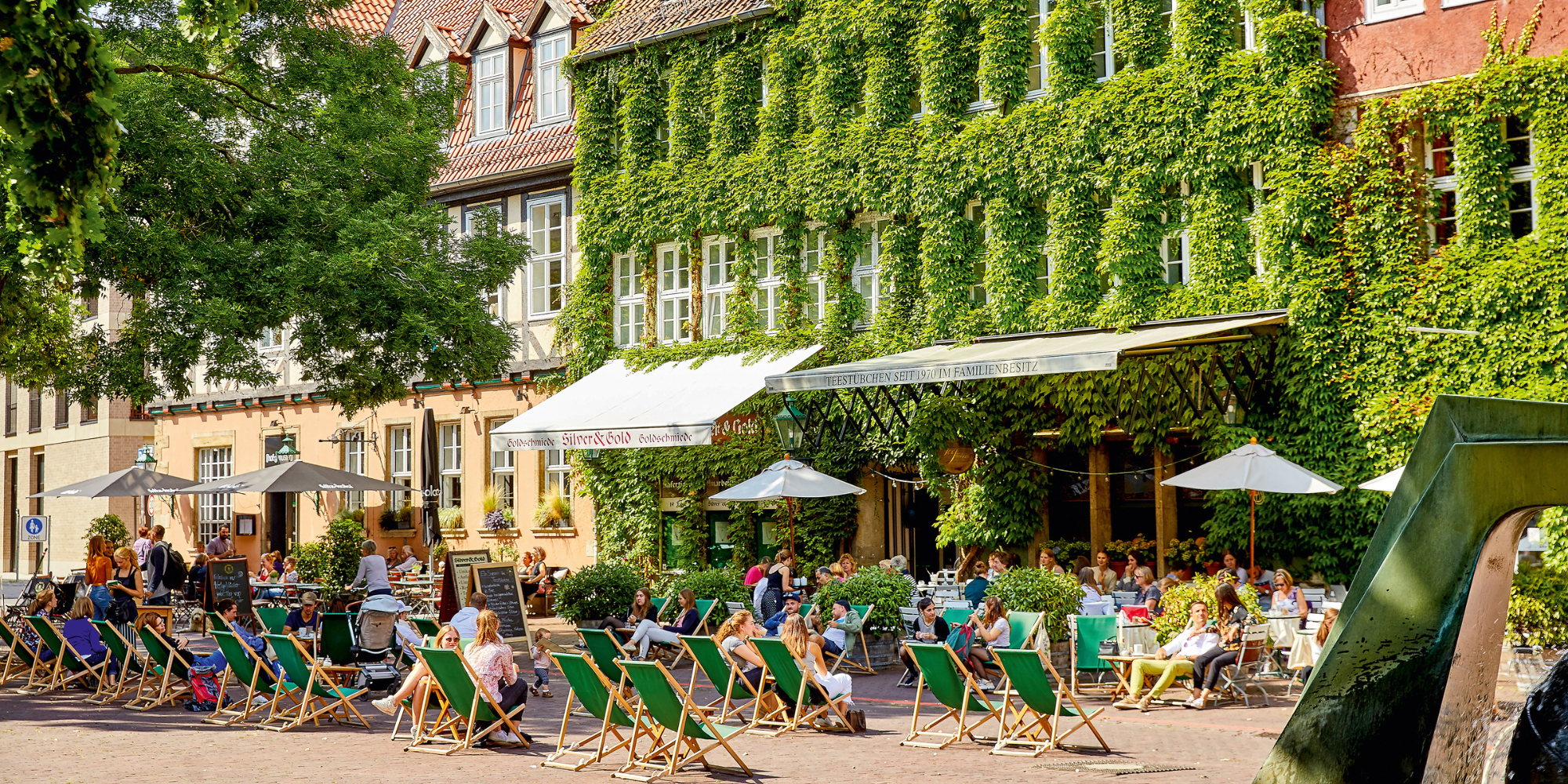 Menschen entspannen in Liegestühlen vor einem begrünten Café in Hannovers Altstadt bei sonnigem Wetter.