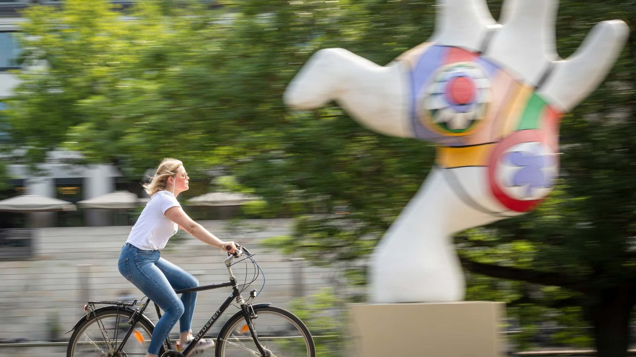 Eine junge Frau fährt mit dem Fahrrad entlang der Nanas am Leibnitzufer.