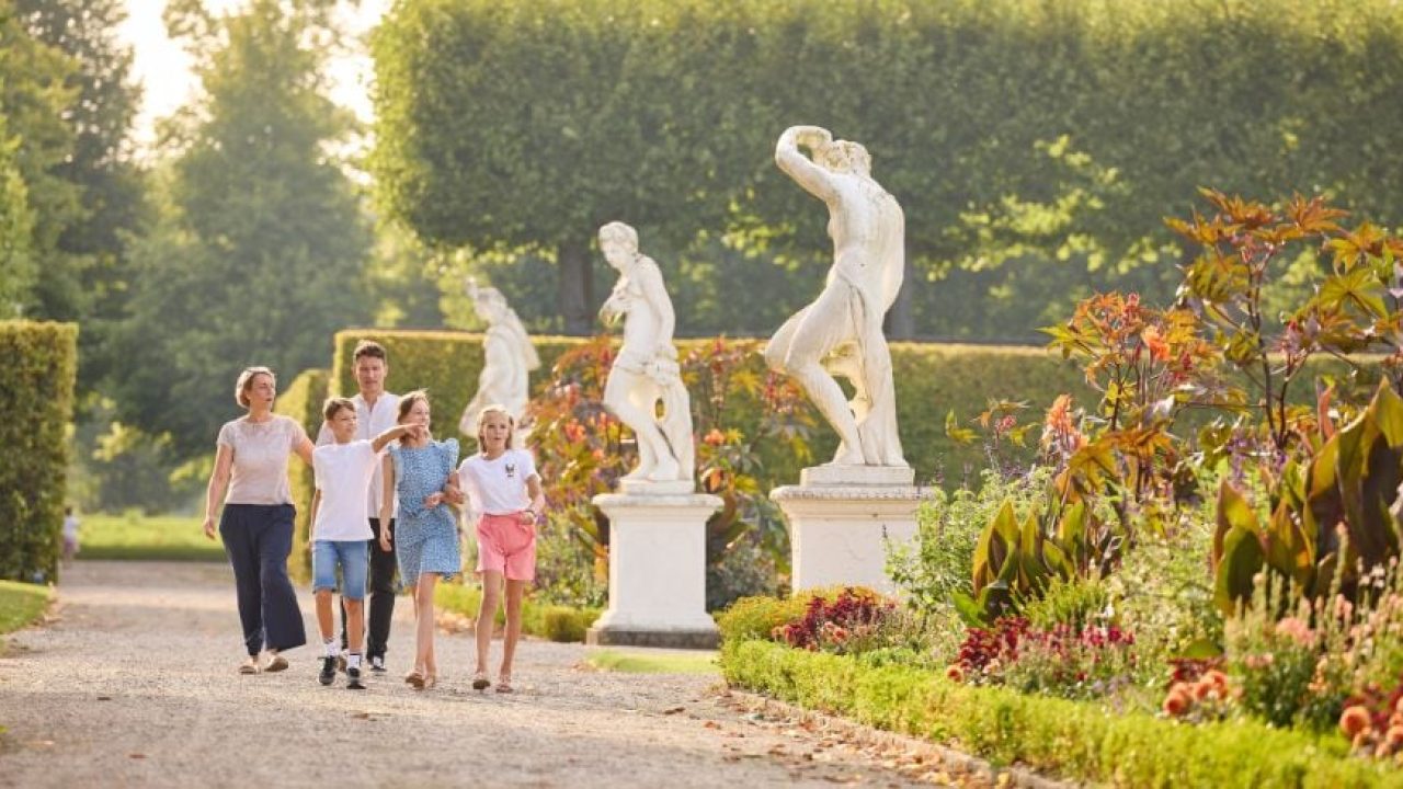 Familie spaziert bei Sonnenschein durch barocken Garten mit Skulpturen und Blumenrabatten.