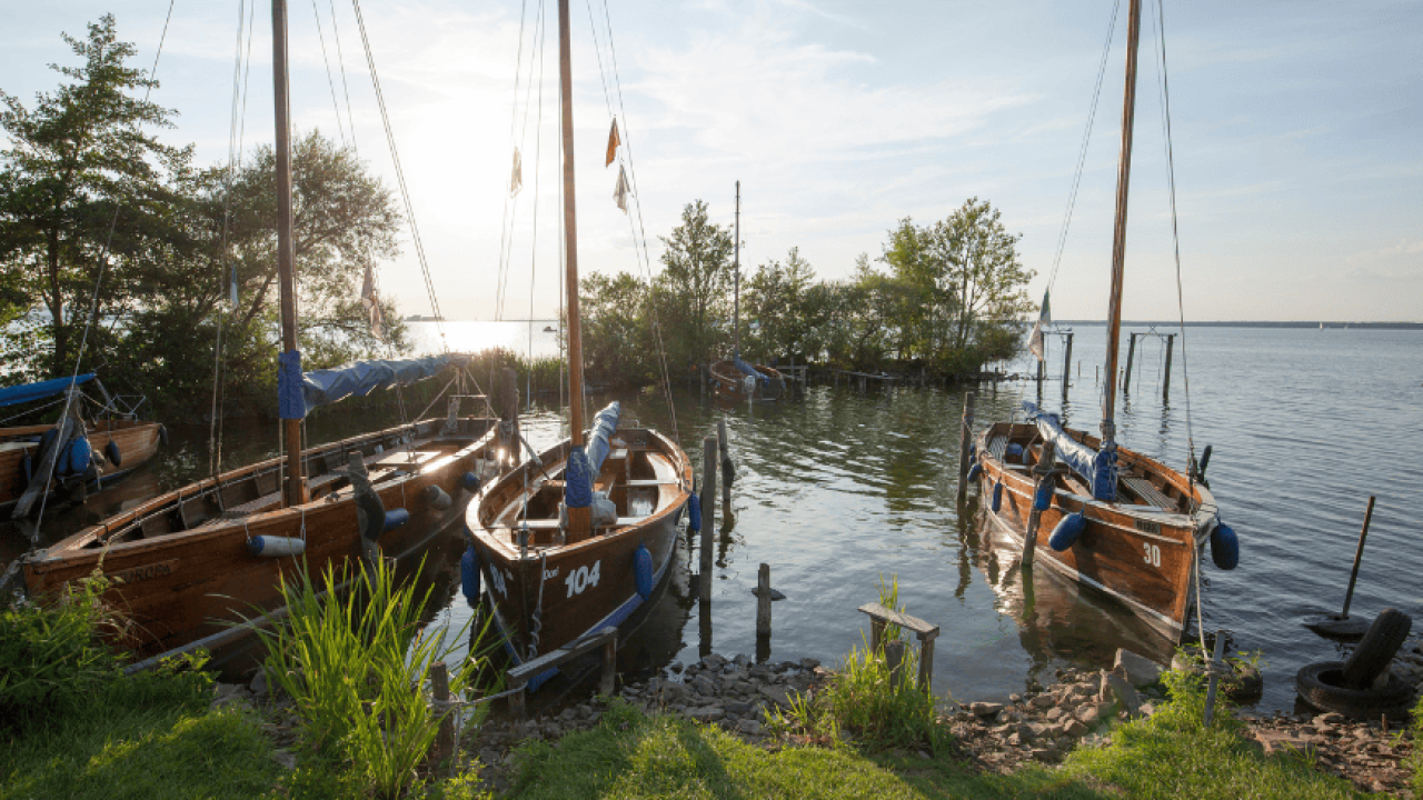 Mehrere Holzsegelboote liegen am Ufer eines ruhigen Sees bei tiefstehender Sonne.