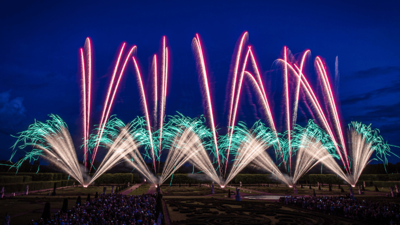 Erlebnispaket: Farbenprächtiges Feuerwerk über barocker Gartenanlage bei Nacht, beobachtet von einer großen Menschenmenge.